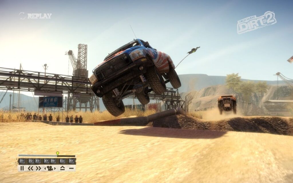 A trophy truck in mid-air over a ramp on a dusty track, cranes and scaffolding behind