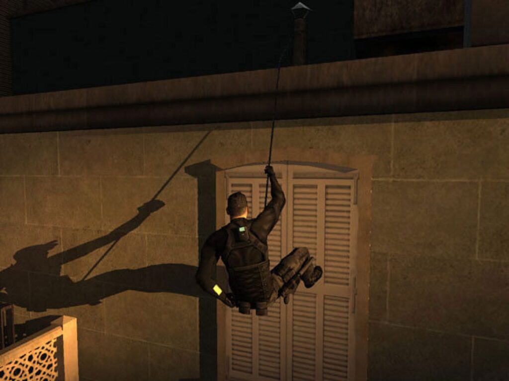 Sam Fisher laat zich aan een touw langs de muur van een huis met luiken zakken, zijn lange schaduw op de muur